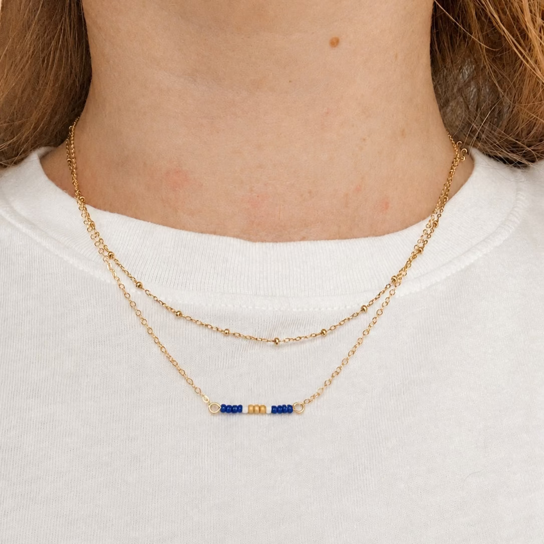 A close-up of a person's neck and upper chest wearing a delicate double-layered gold necklace with a small central bar featuring blue and gold beads, styled with a white crewneck shirt.