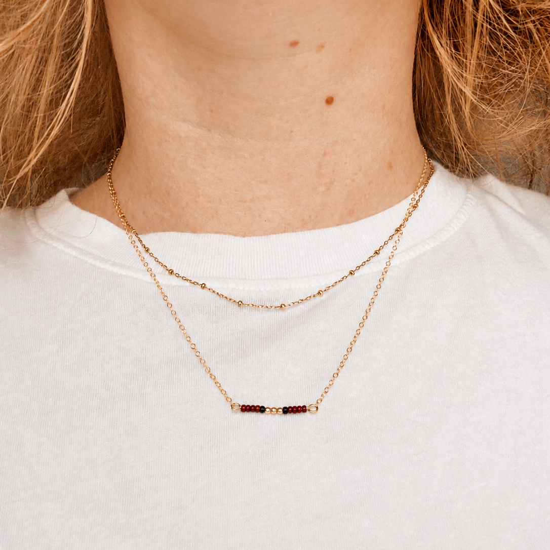 A close-up of a model's neck and upper chest wearing a delicate layered necklace. The necklace features a gold chain with a small horizontal bar set with dark red, black and gold filled beads, layered over a thinner gold chain.