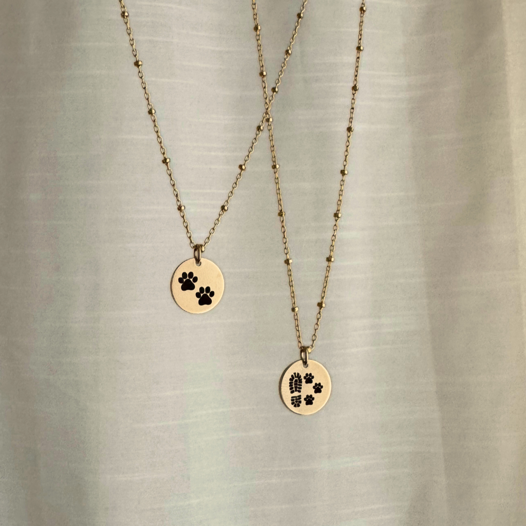 Two gold necklaces with paw print pendants on a textured beige background