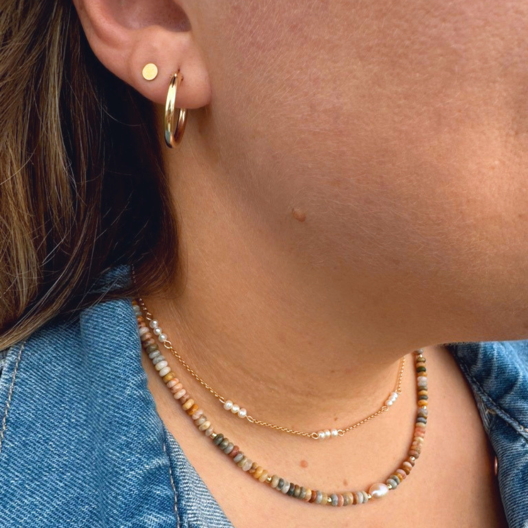 Close-up of a person wearing layered necklaces and earrings, including a gold hoop earring, a small gold disc stud, and two delicate beaded necklaces over a blue denim top.