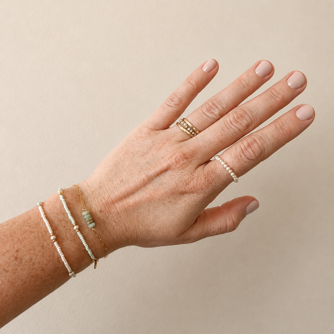 A close-up of a person's hand and wrist wearing delicate jewelry, including a gold ring with a green stone, a silver beaded ring, and several thin silver bracelets, against a plain beige background.