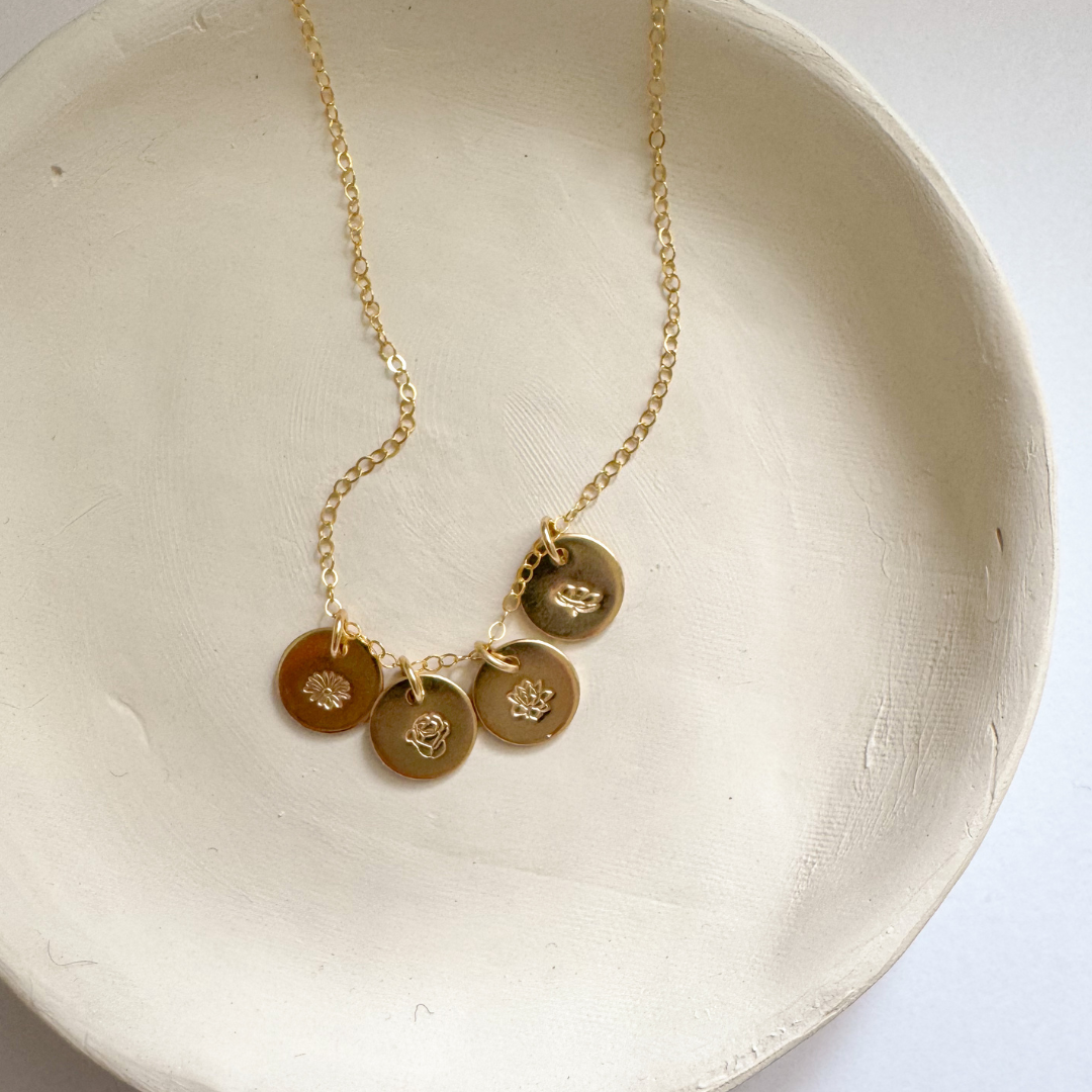 A delicate gold chain necklace with four small round gold charms, displayed on a textured off-white ceramic dish against a light background.