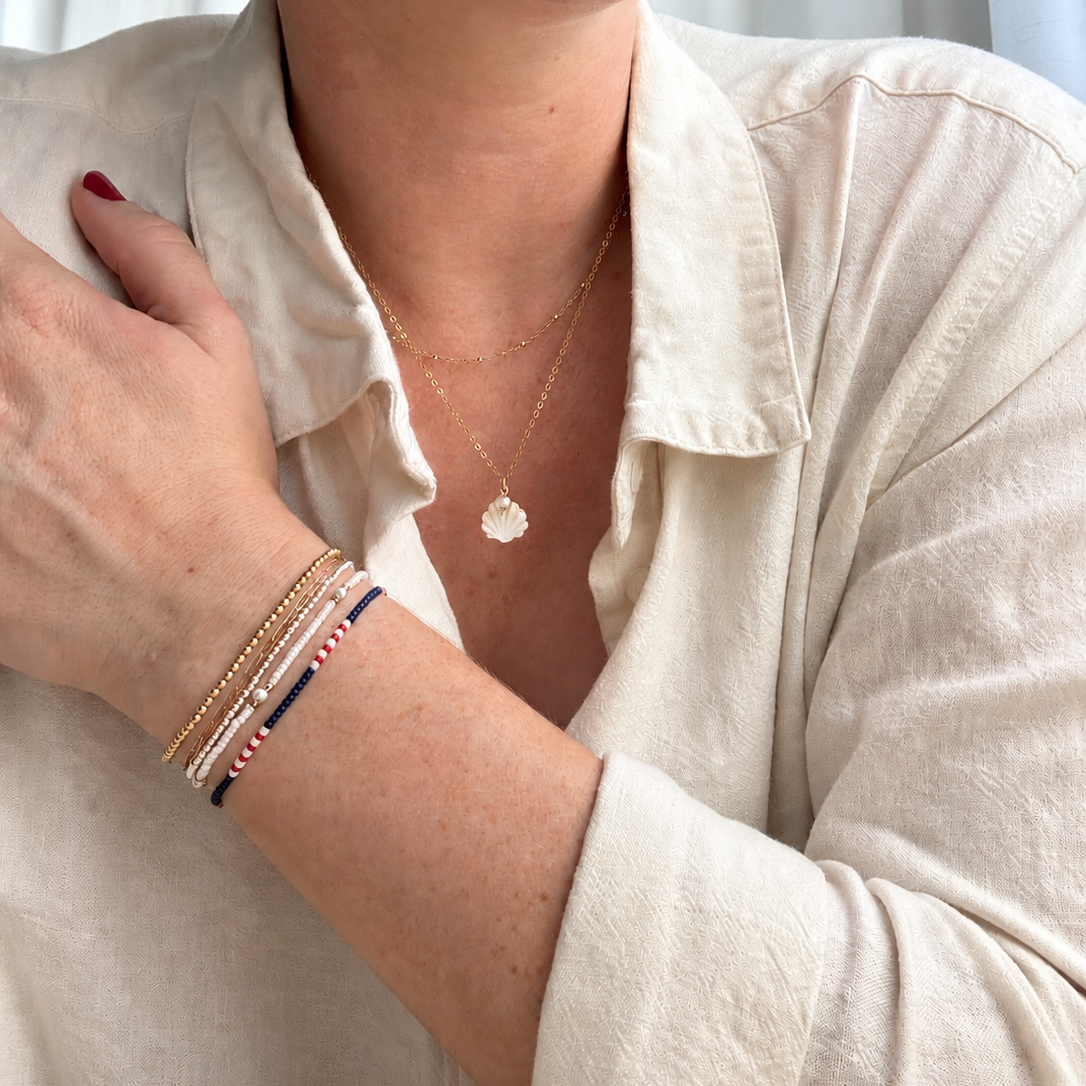 A close-up of a person wearing a delicate gold necklace with a small round pendant and several stacked bracelets on one wrist, including a navy and red beaded bracelet, against a light neutral background.