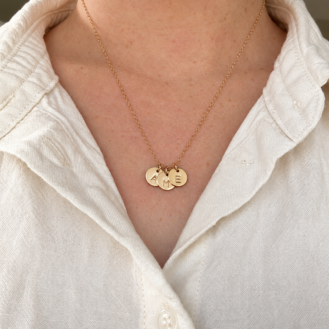 A close-up of a person wearing a delicate gold chain necklace with three small round gold pendants, styled with a white linen button-up shirt.