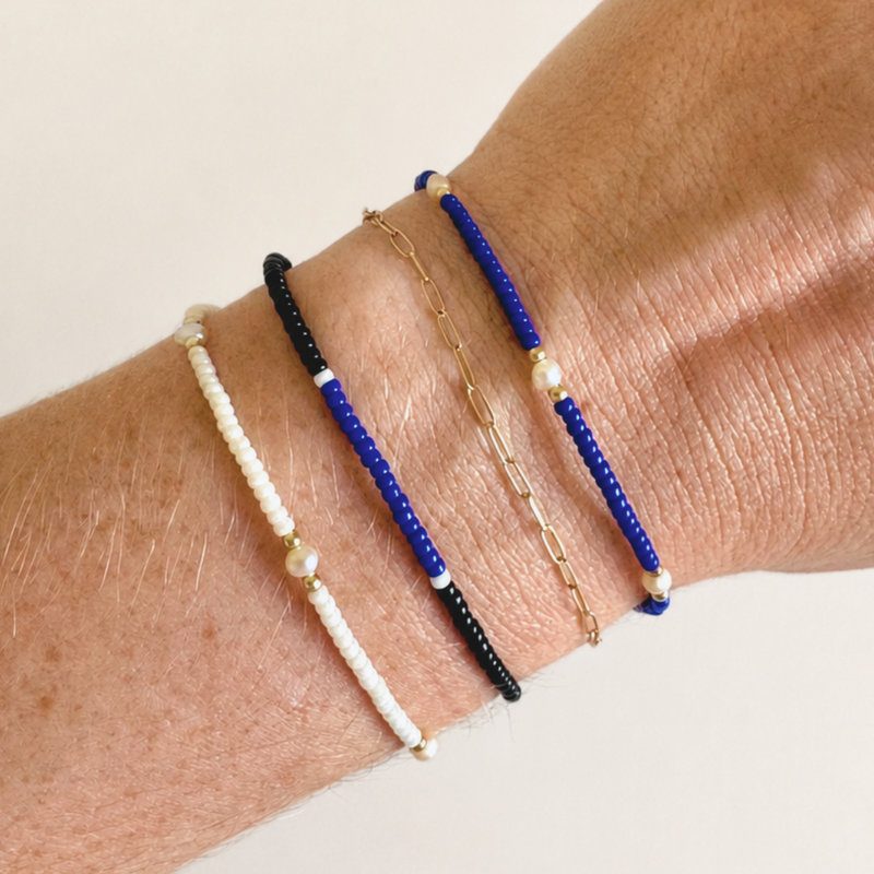 Four delicate beaded and chain bracelets displayed on a person's wrist against a plain light beige background.