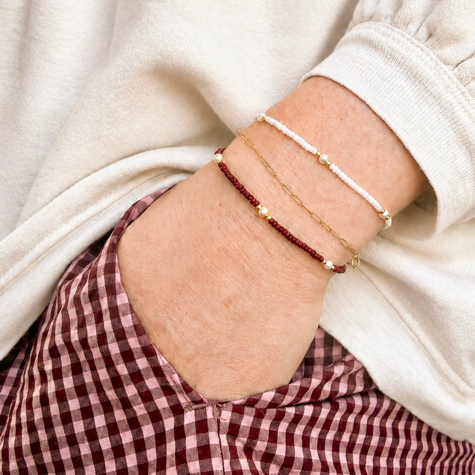 Three delicate bracelets worn on a person's wrist, featuring white beads, a gold chain with small gold beads, and a dark red beaded bracelet with gold star accents, styled with a cream sweatshirt and a red-and-white gingham skirt.
