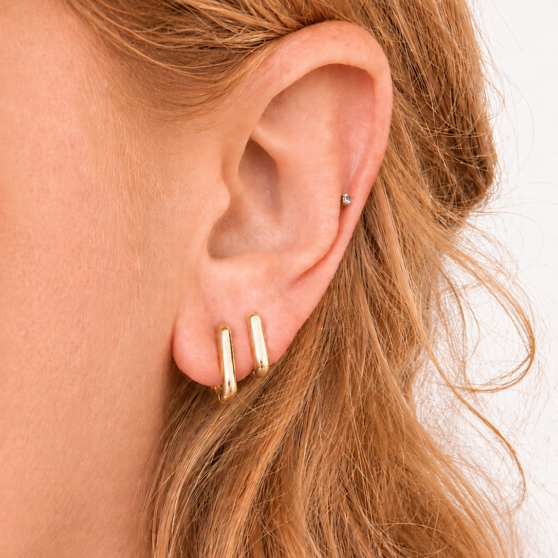 A close-up of a person's ear wearing two slim gold hoop earrings on the lobe and a small silver stud on the upper ear cartilage, with light brown hair against a plain light background.