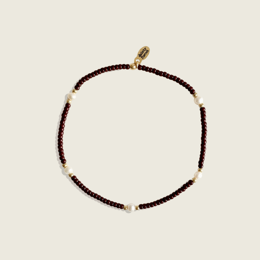 A beaded necklace featuring dark brown faceted beads, small white pearl accents, and gold spacer beads, with a gold clasp displaying the brand name 'SARAH MARSHALL'. The necklace is photographed on a solid, warm off-white background.