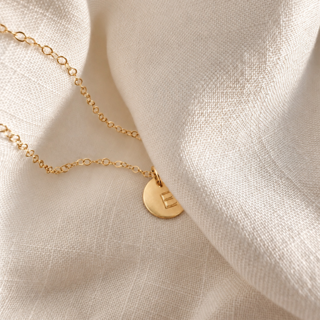 A delicate gold chain necklace with a small round gold pendant engraved with the letter 'E', resting on draped cream-colored linen fabric.