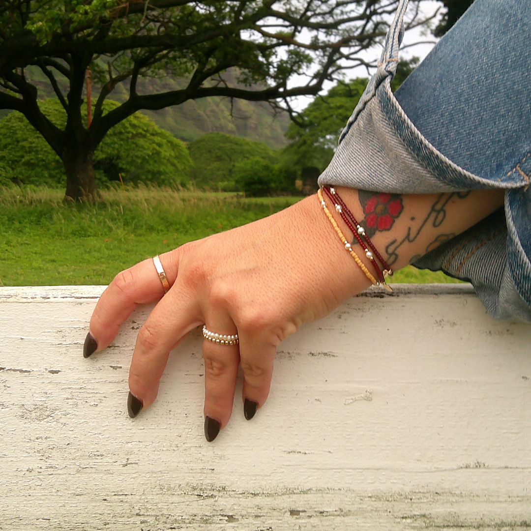 A close-up of a person's hand resting on a white wooden fence in an outdoor park setting, wearing silver rings and beaded bracelets.
