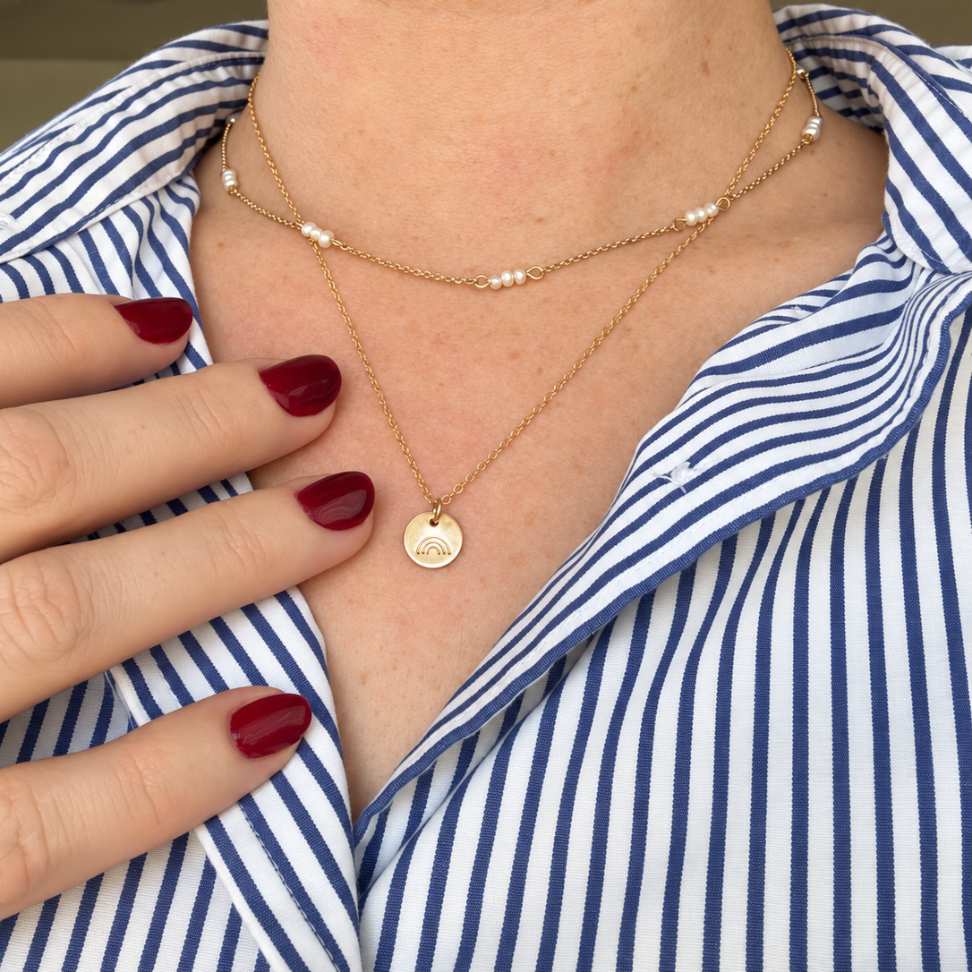 A close-up of a person wearing layered gold necklaces, including one with small white pearl-like beads and a round gold pendant, over a blue and white striped collared shirt.