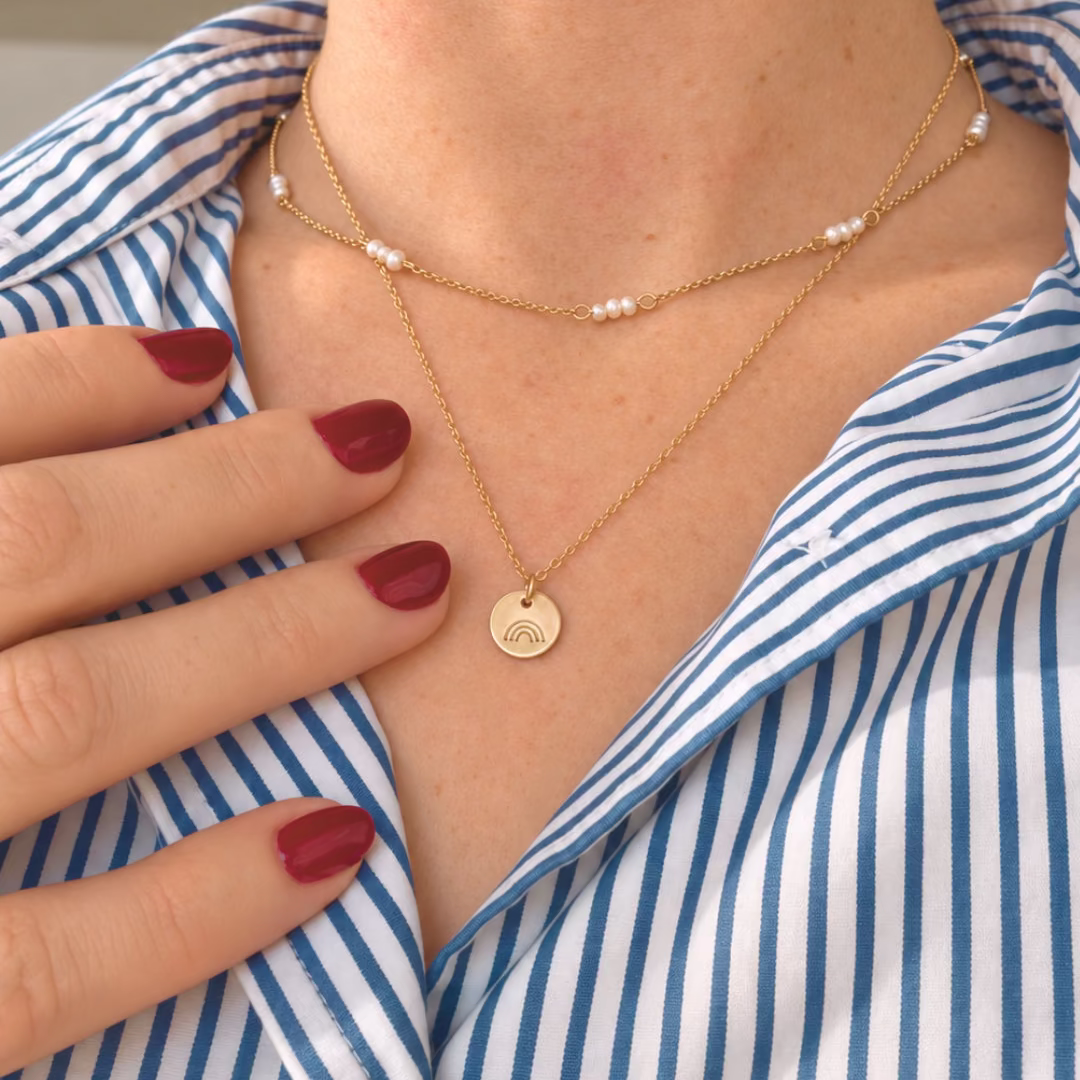 A close-up of a person wearing layered gold necklaces, including a delicate chain with small white pearl beads and a round gold pendant, paired with a blue and white striped shirt.