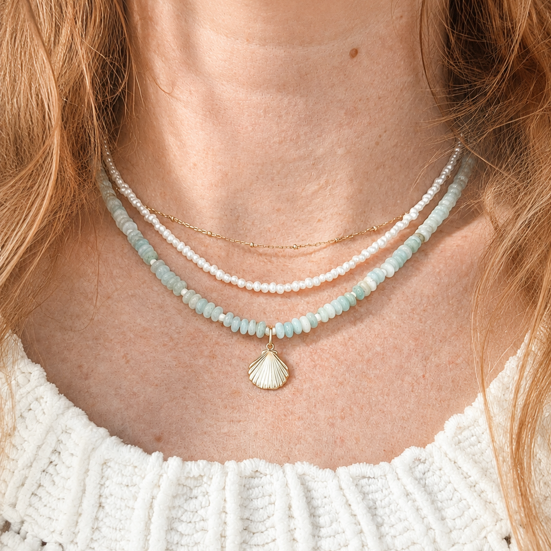 A close-up of a model wearing a layered necklace set. The set includes a delicate gold chain, a strand of white pearls, and a strand of light blue beads with a small gold seashell pendant.