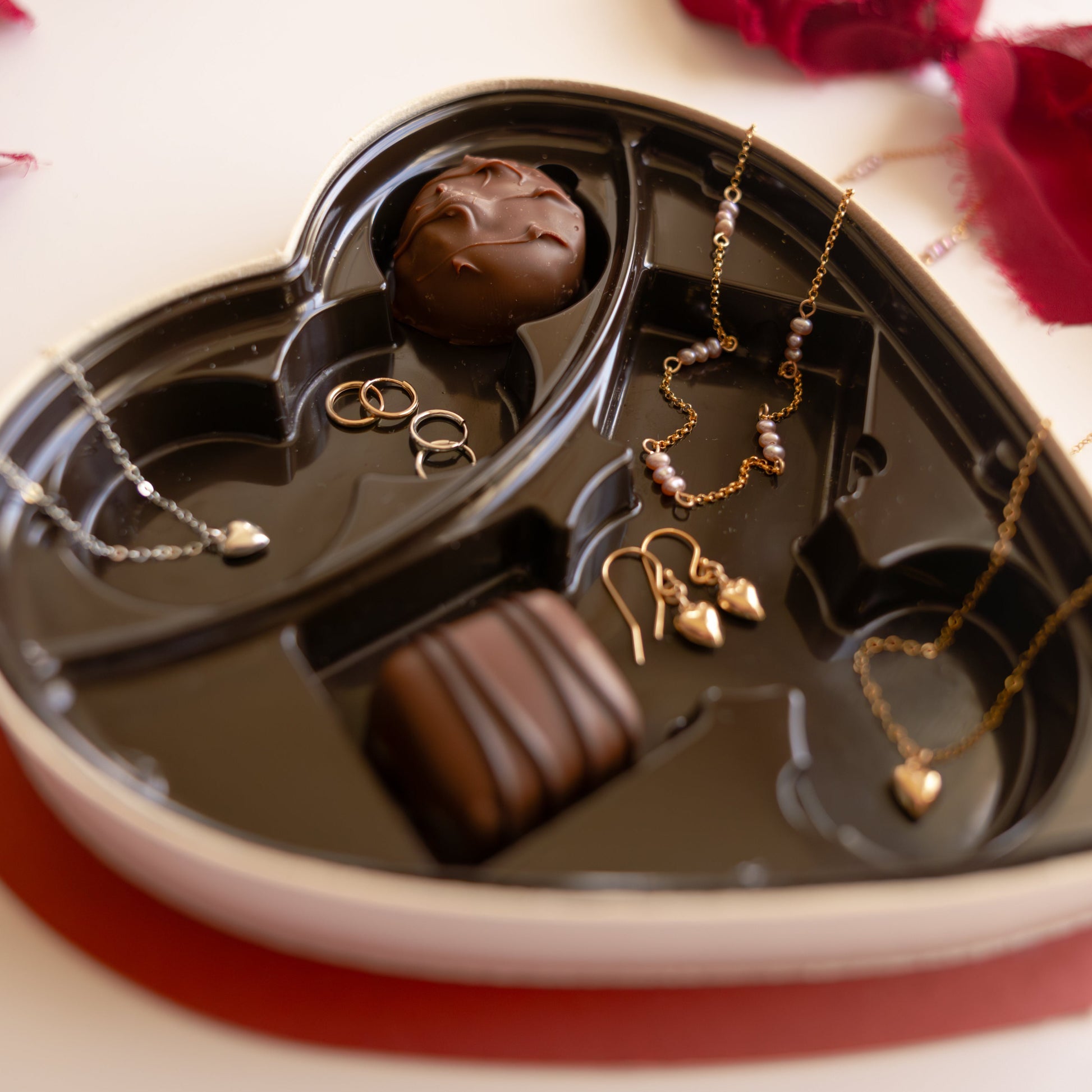 Heart-shaped chocolate tray with jewelry and chocolates on a white surface with red rose petals.