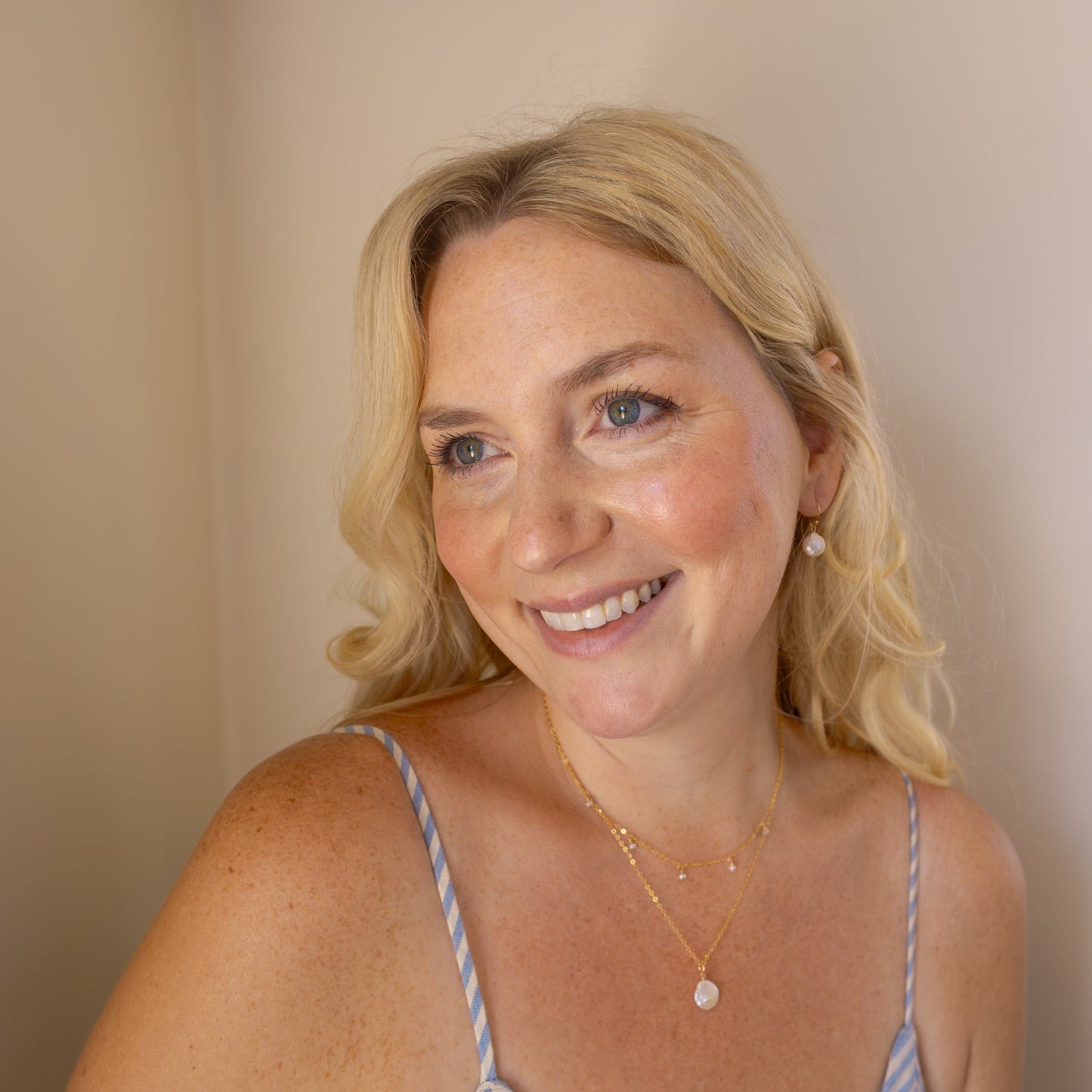 Woman wearing a blue and white striped dress against a beige background with the coastal coin earrings, timeless perl necklace, and coastal coin necklace