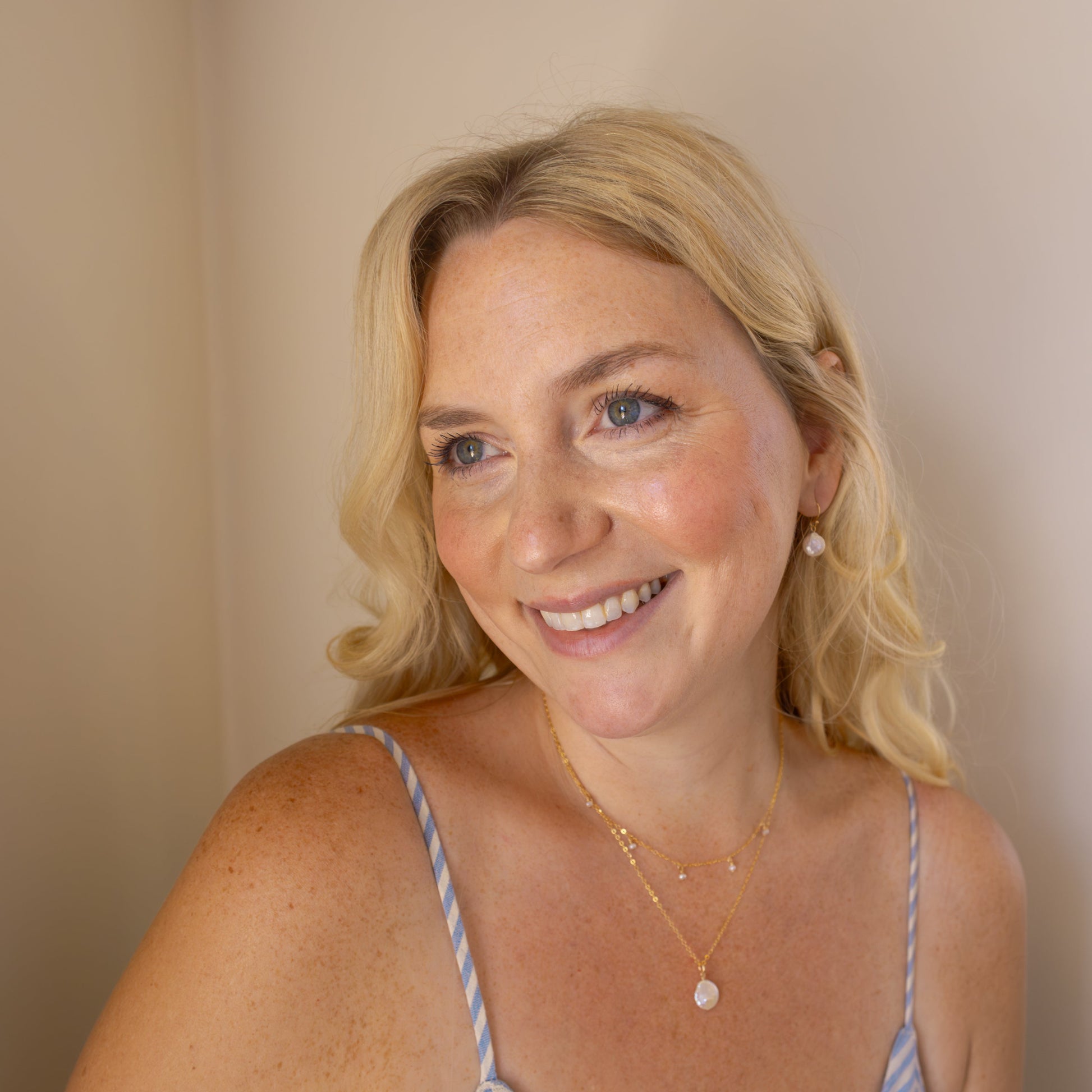 Woman wearing a blue and white striped dress against a beige background with the coastal coin earrings, timeless perl necklace, and coastal coin necklace