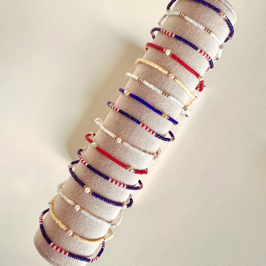 A set of beaded friendship bracelets in red, white, and blue, displayed on a beige fabric roll-up stand against a plain off-white background.