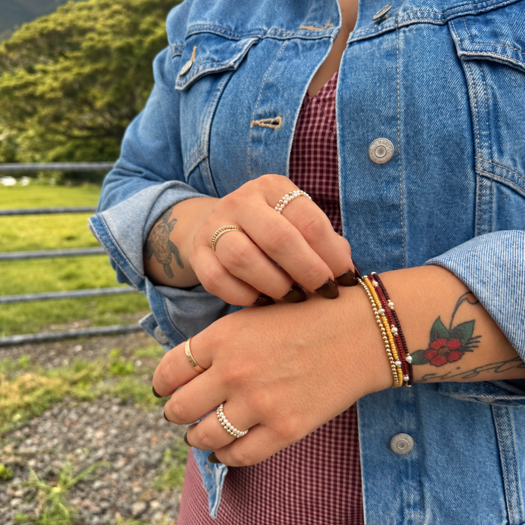 A close-up of a person outdoors wearing multiple bracelets on their wrist, including a dark brown beaded bracelet and a red and gold beaded bracelet, styled with a blue denim jacket and a red gingham top against a blurred mountain background.