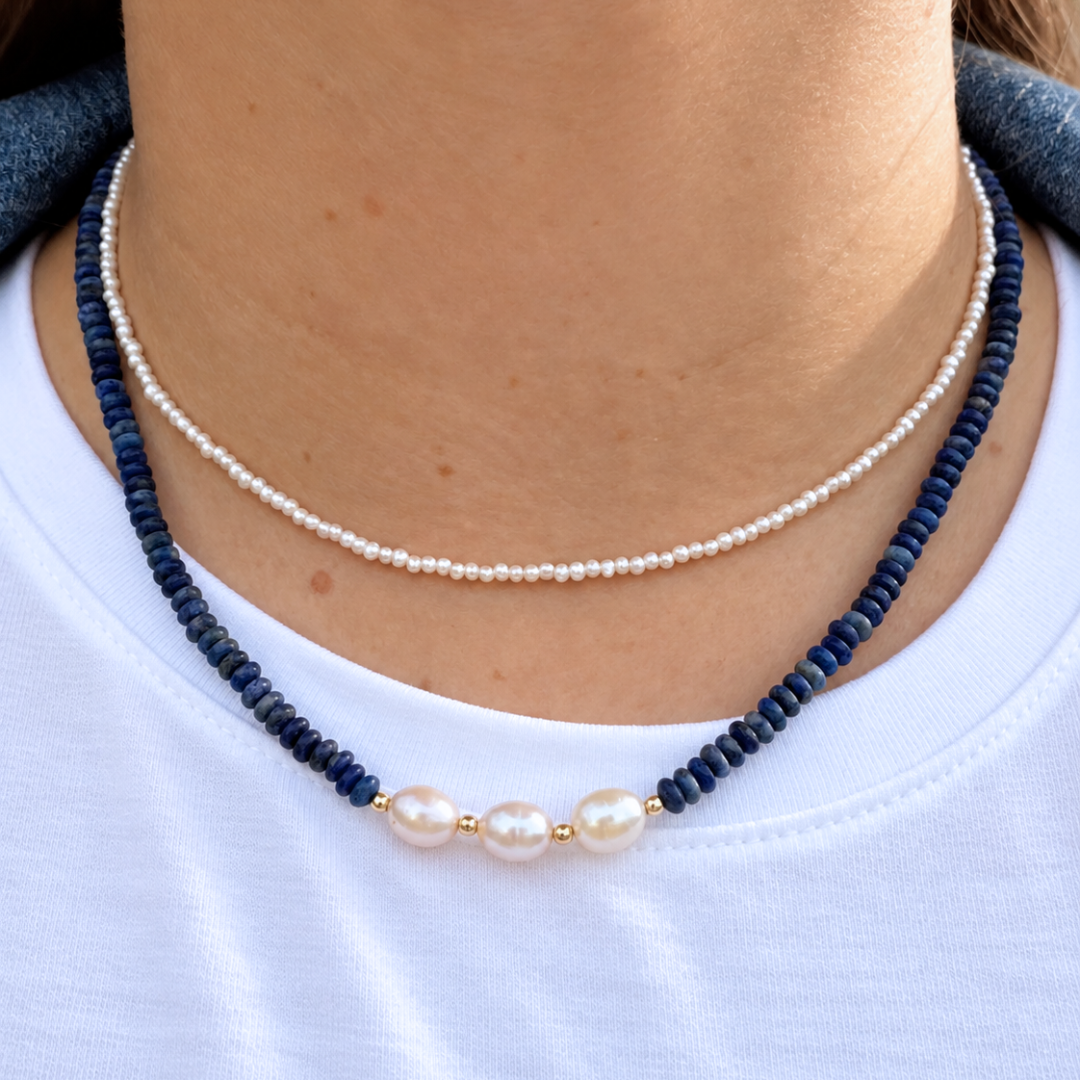 A close-up of a person wearing a double-strand necklace. The top strand is made of small white pearls, and the bottom strand is made of dark navy blue beads with three larger white pearls at the center.