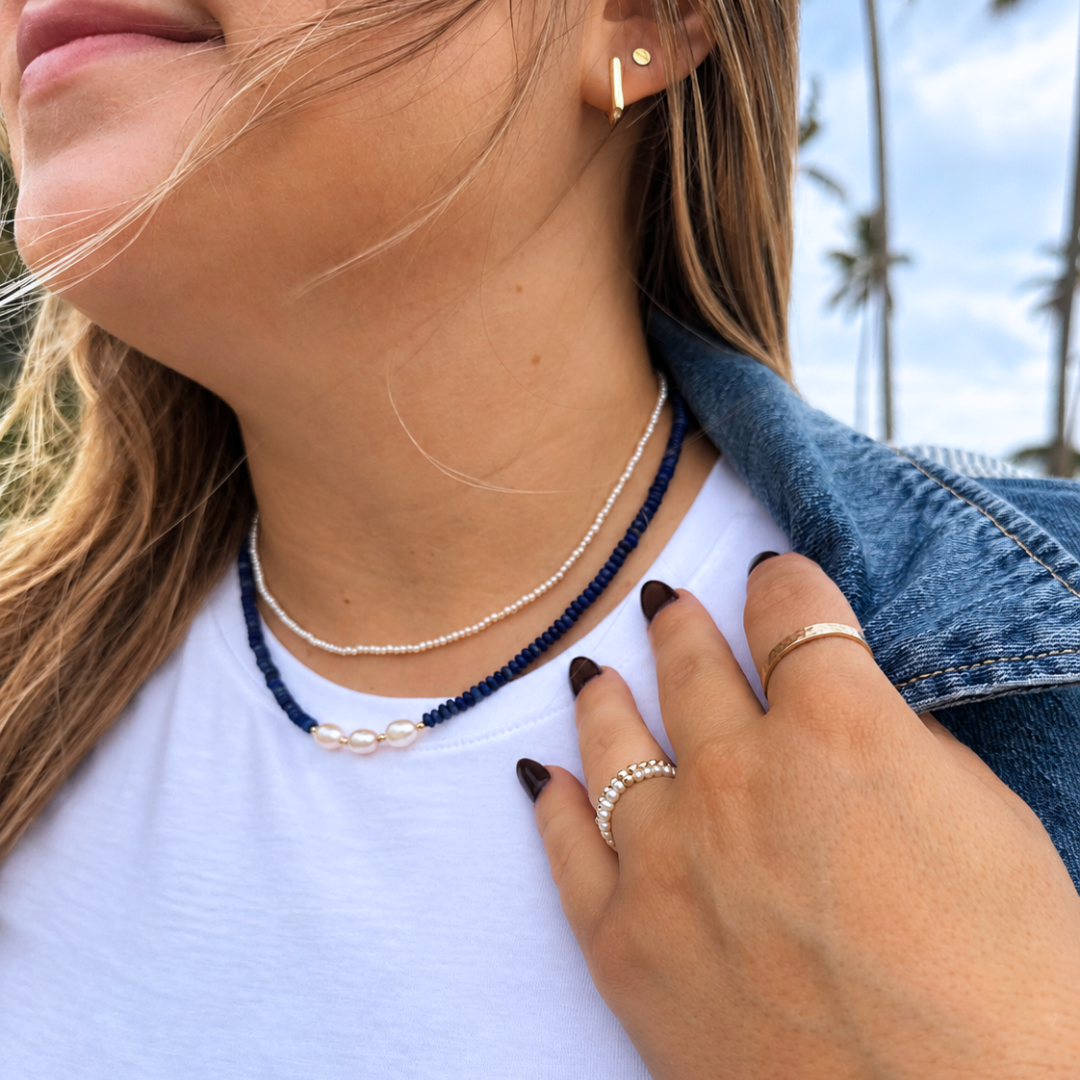 A close-up lifestyle shot of a woman wearing layered necklaces, including a navy blue beaded necklace with a white pearl accent and a silver beaded necklace, along with rings and a denim jacket.