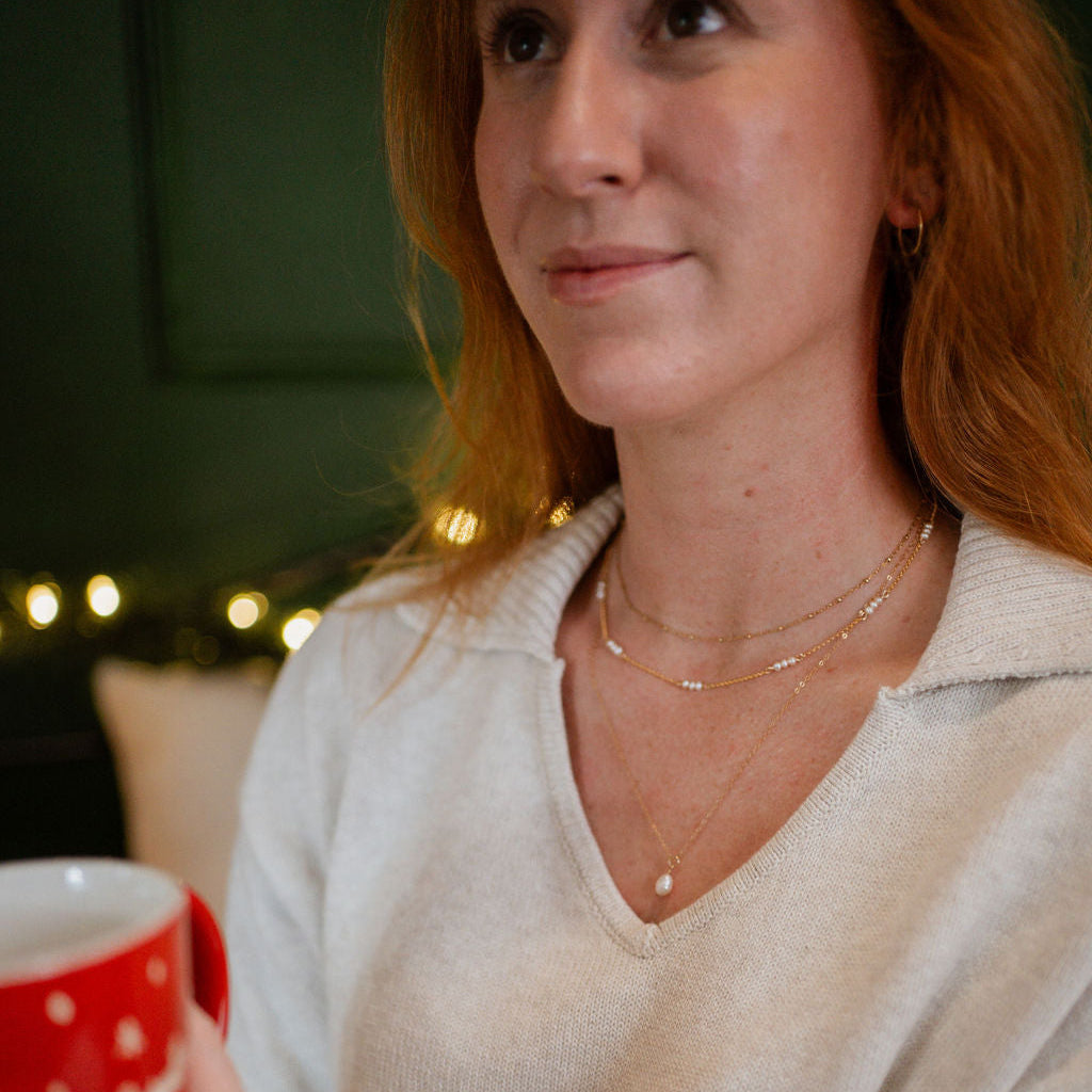Woman holding a red mug with white polka dots, wearing a beige sweater and gold jewelry.