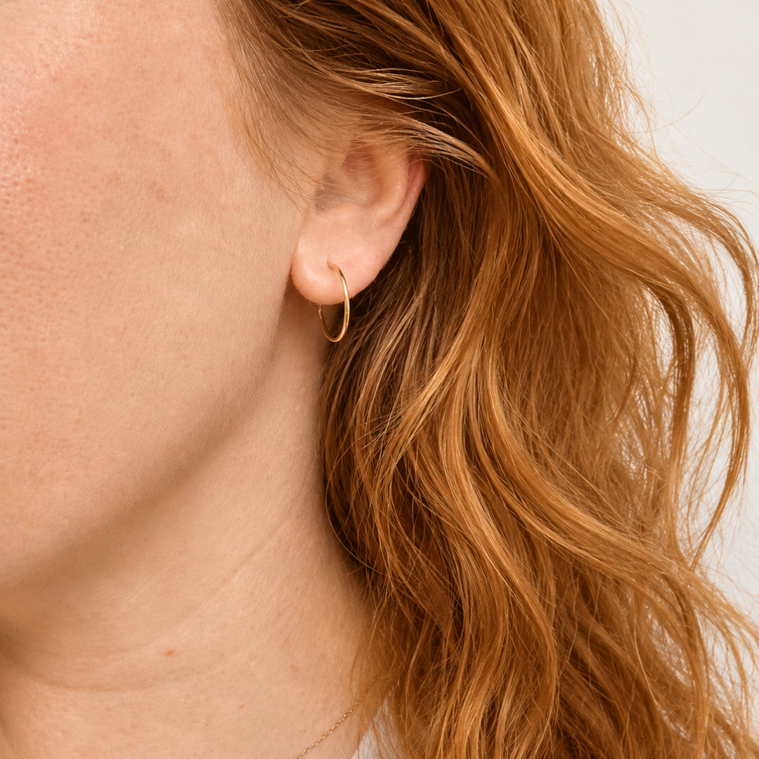 A close-up of a person's ear wearing a delicate gold hoop earring, with wavy red hair and a thin gold chain necklace visible against a plain light background.
