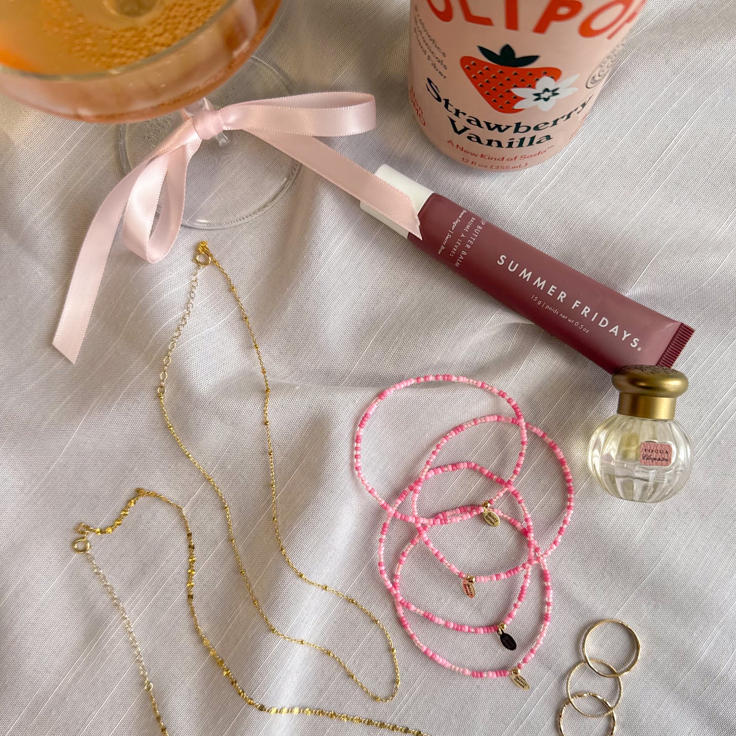 girly pop pink bracelets with flay lay of necklaces, rings, on a white background with drinks, perfume, and lipstick