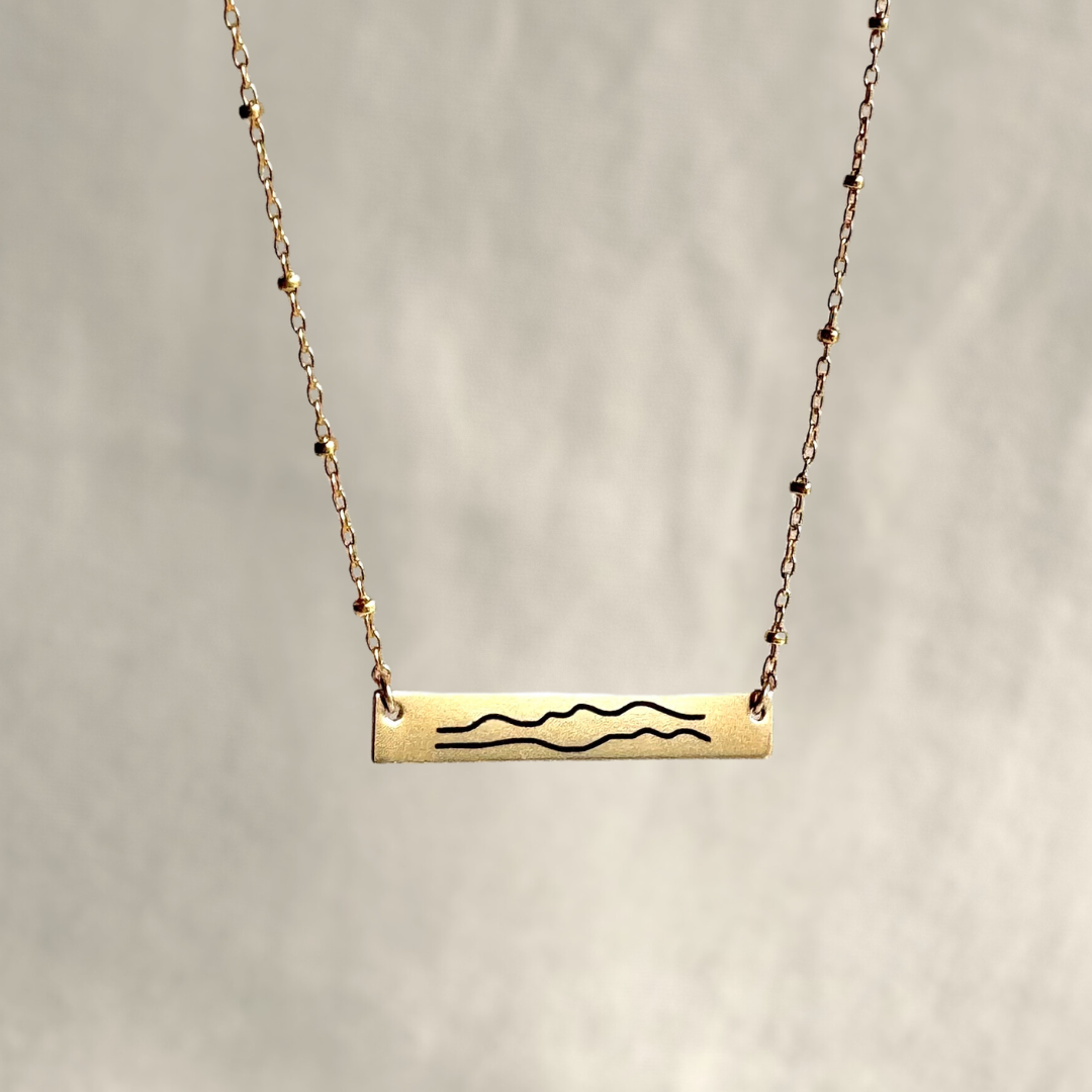 Gold necklace with a bar pendant featuring wavy lines on a neutral background