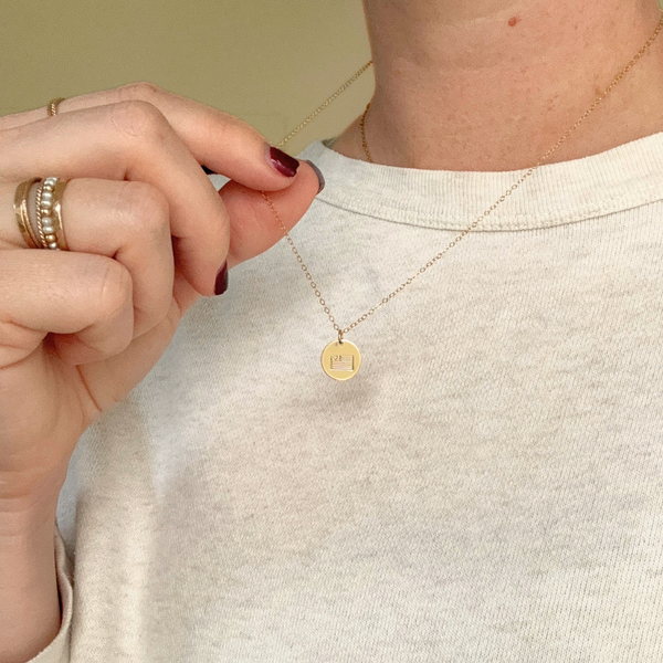 Person wearing a gold necklace with a round pendant, holding it near their chest.
